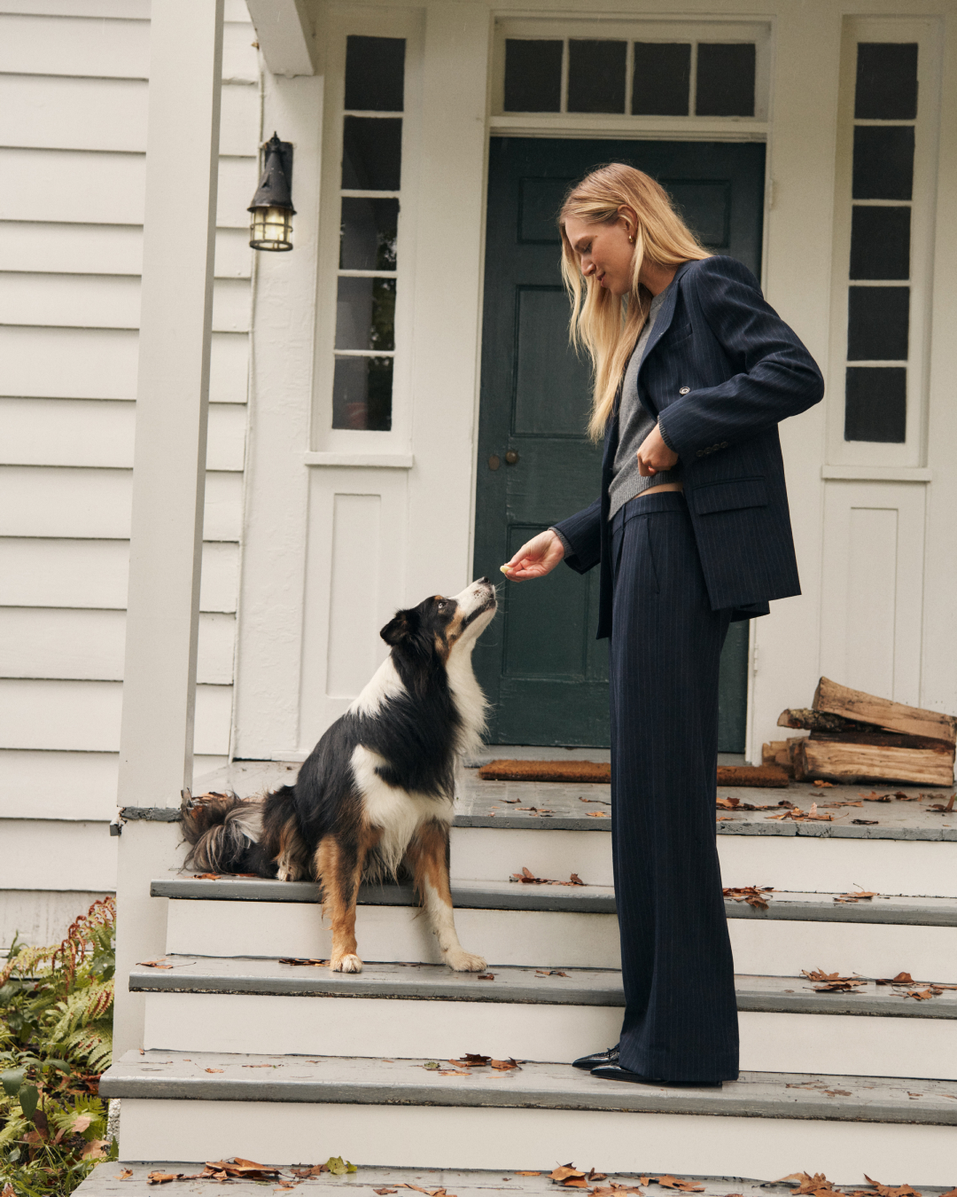 A woman wearing a navy blue suit, standing on white stairs, feeding a treat to a tri-colored dog