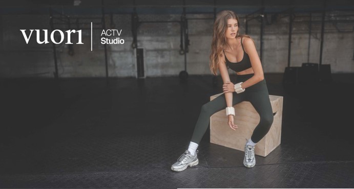 A girl in green leggings and a sports bra, sitting on a wooden cube in a pilates studio