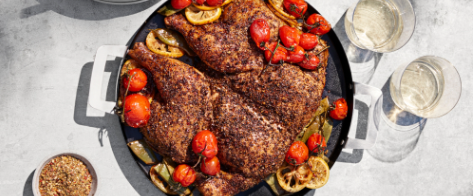 A pan of seasoned chicken with tomatoes and lemon on a white marble counter