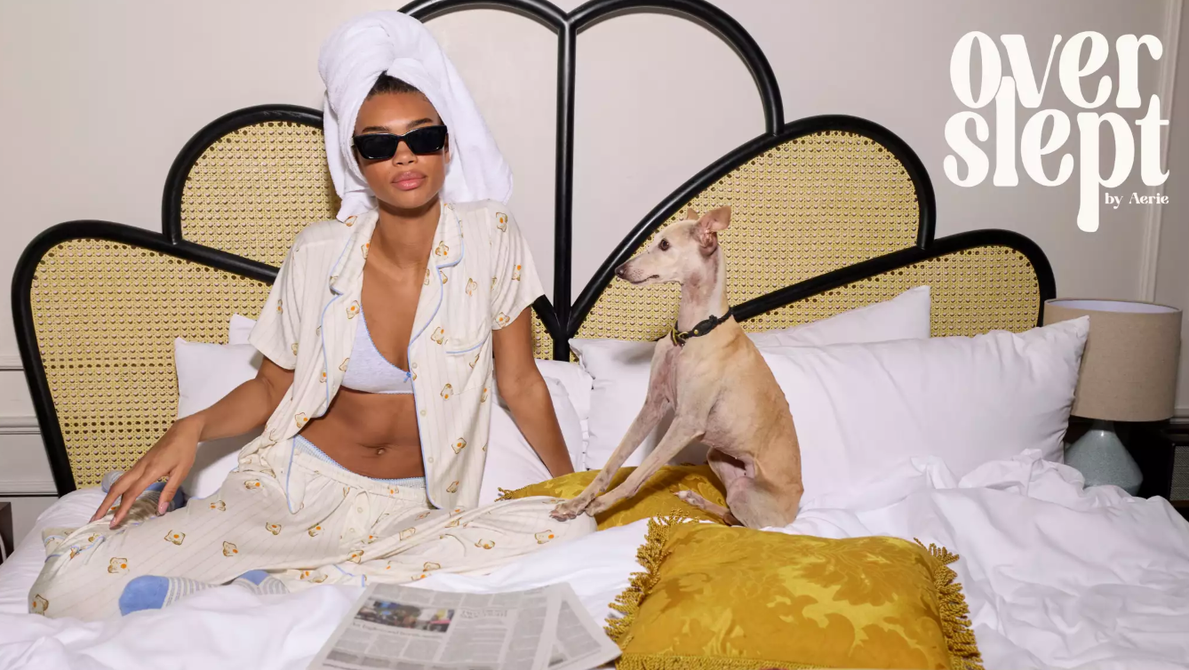 A young woman sitting on a bed in her pajamas with a white dog next to her reading the newspaper