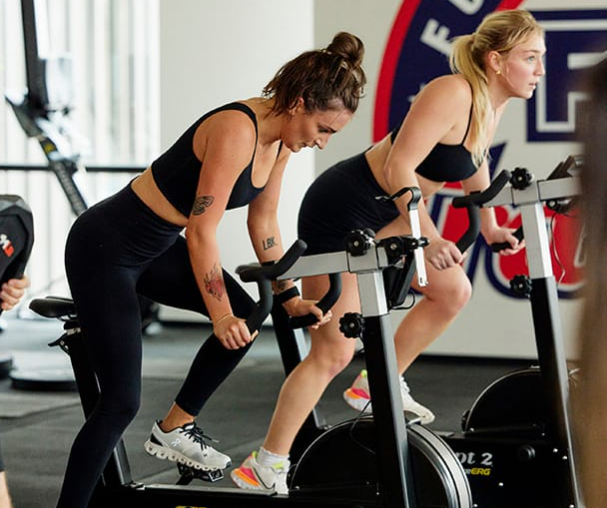 Two women in workout gear riding on stationery bikes in a gym