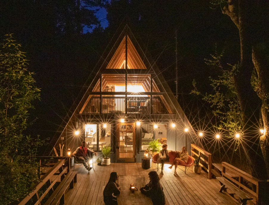 People sitting on the deck of an A frame house at night. There is furniture on the deck and string lights