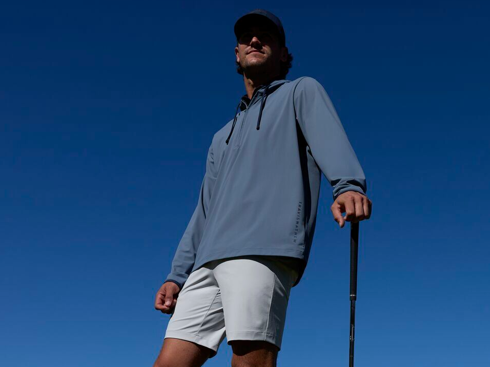 A young man in a baseball cap, shorts and zip jacket standing with a golf club under his left hand