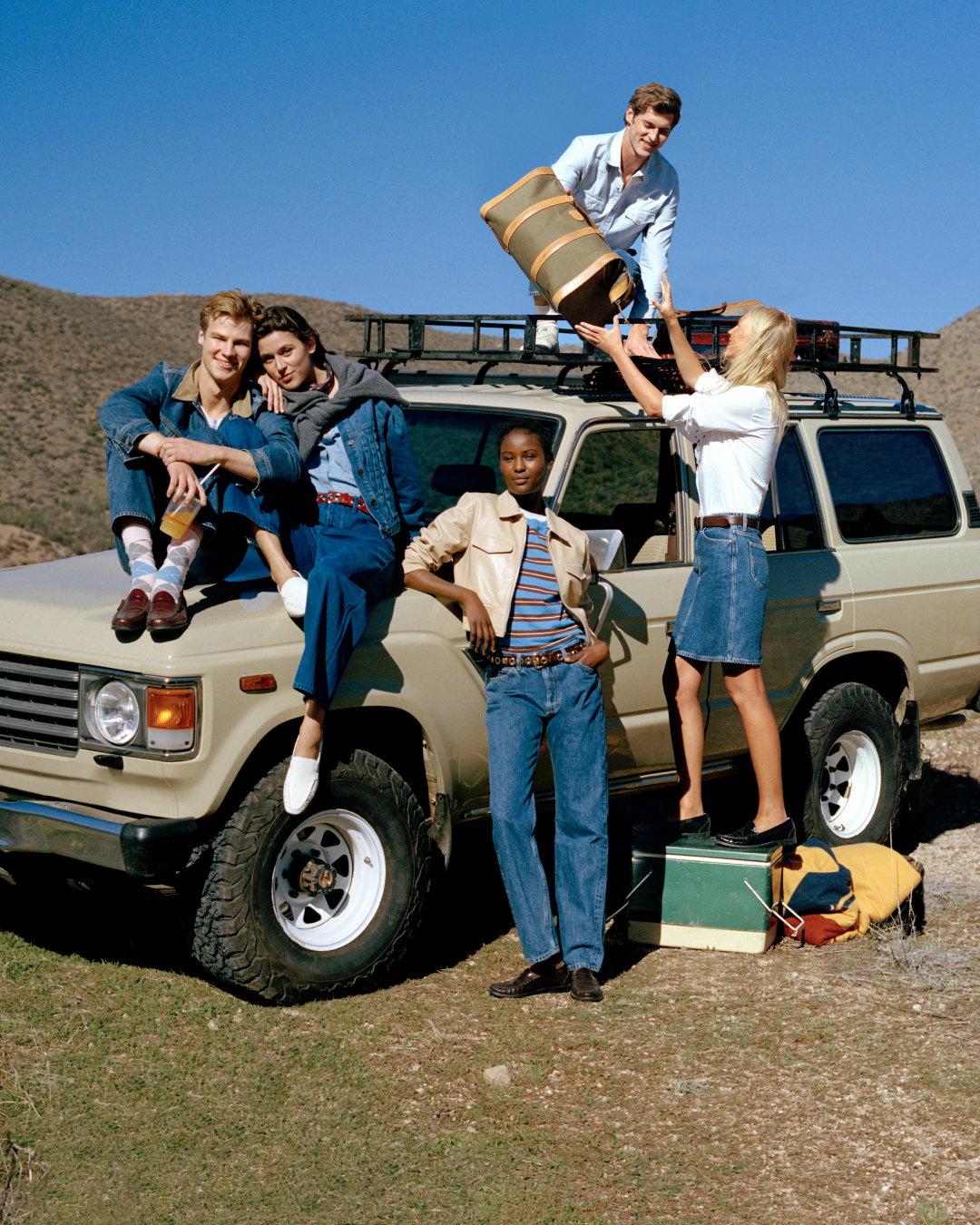 A group of young men and women in denim loading luggage onto a beige jeep