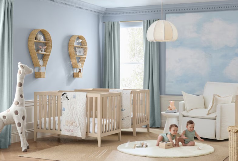 Two babies on a round white carpet in a baby's room with blue walls, two beige cribs, wall decor and a tall stuffed giraffe