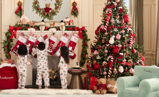 Three kids in matching pajamas reaching for their holiday stockings hanging on the fireplace in a decorated living room with a large Christmas tree