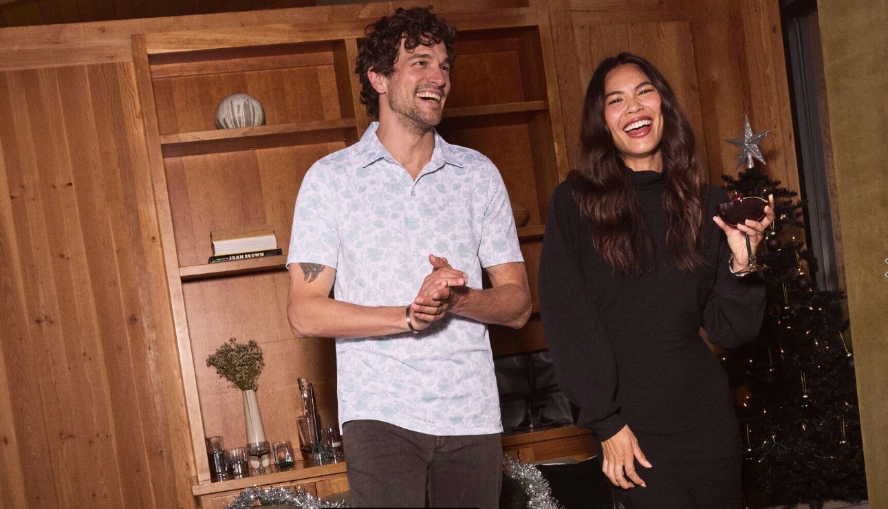 A young couple standing in front of a bookshelf. The woman is wearing a black dress holding a glass of wine, the man is wearing a short sleeve shirt and pants