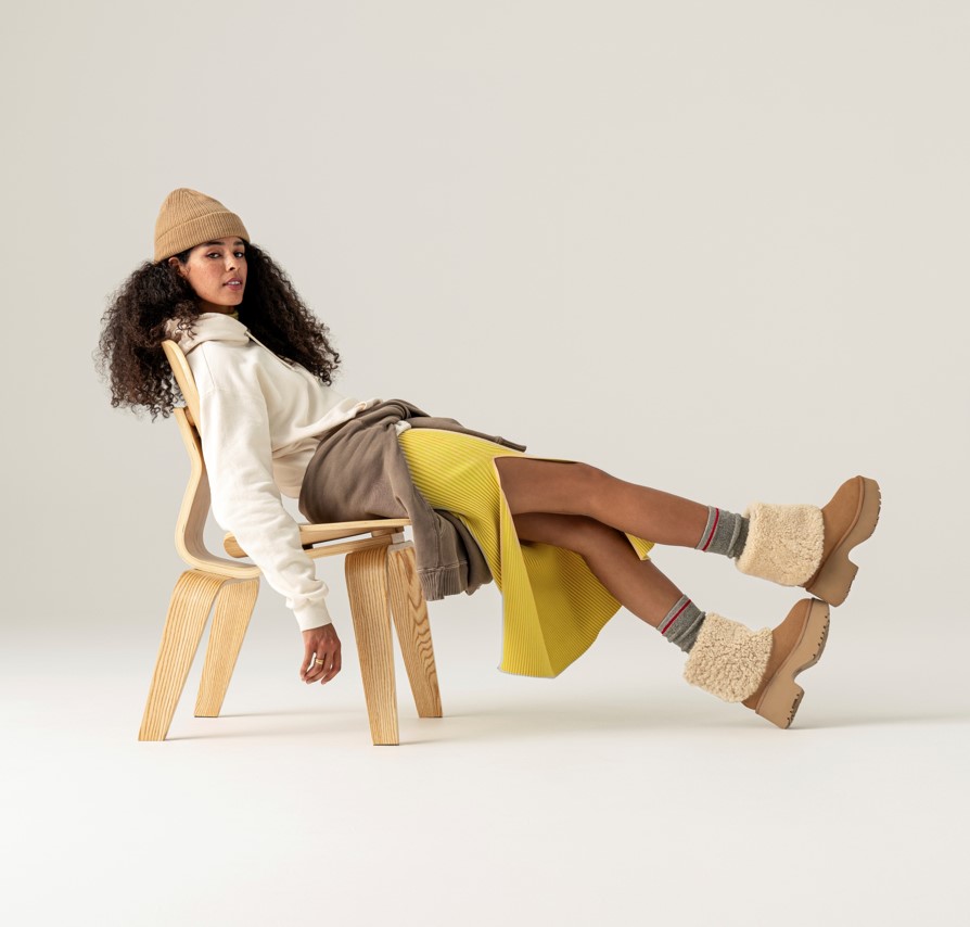 A woman slouching on a wooden chair wearing a cream sweater, and yellow skirt with uggs