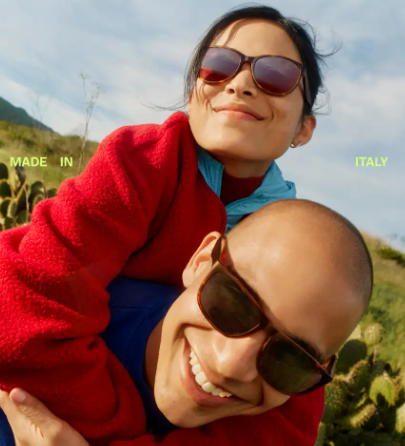 A woman in a red jacket on the shoulders of a man. Both are wearing Warby Parker sunglasses