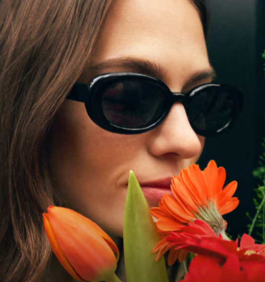 A woman wearing black sunglasses smelling an orange flower 