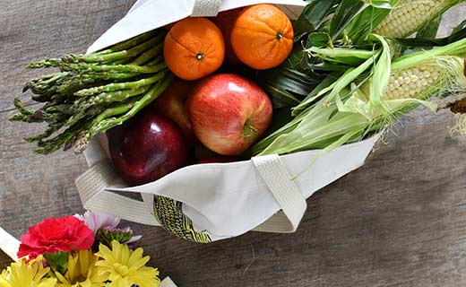 White tote bag with red apples, oranges, corn on the cob, asparagus, and daisies 