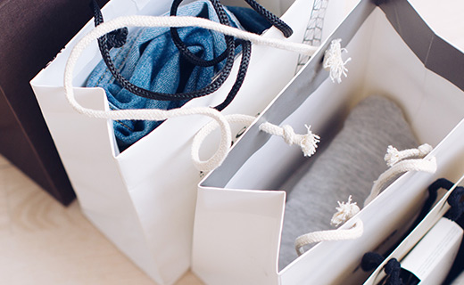 Two white paper shopping bags showing a pair of jeans folded in the bag on the left and grey sweater folded in the bag on the right.