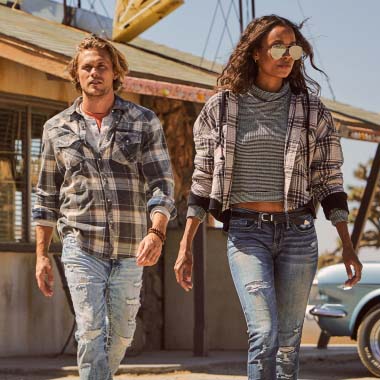 A man wearing ripped jeans with a plaid shirt, a woman wearing ripped jeans, a striped shirt, and a plaid jacket.