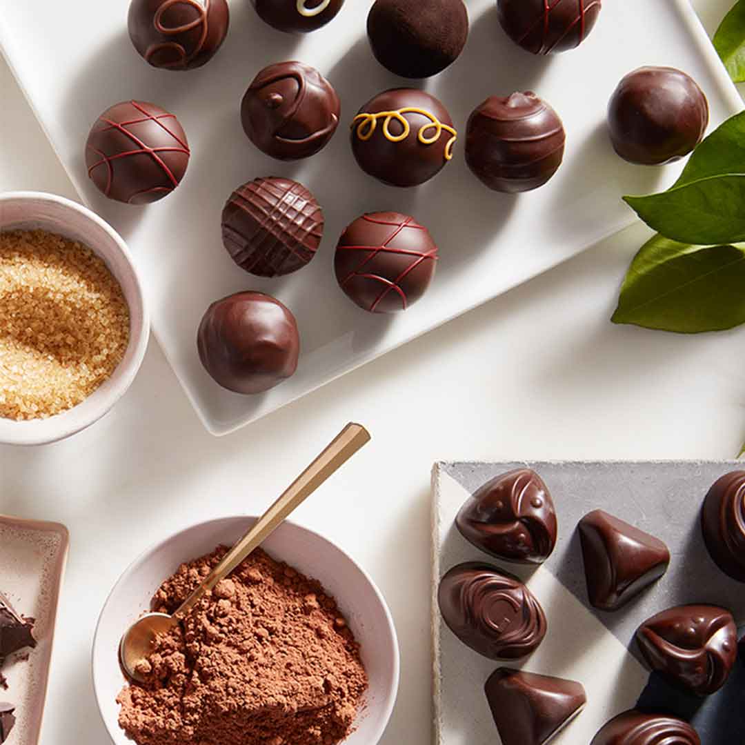 Dark chocolate truffles and dark chocolate candies displayed on a plate with a bowl of cocoa along with a bowl of raw sugar.