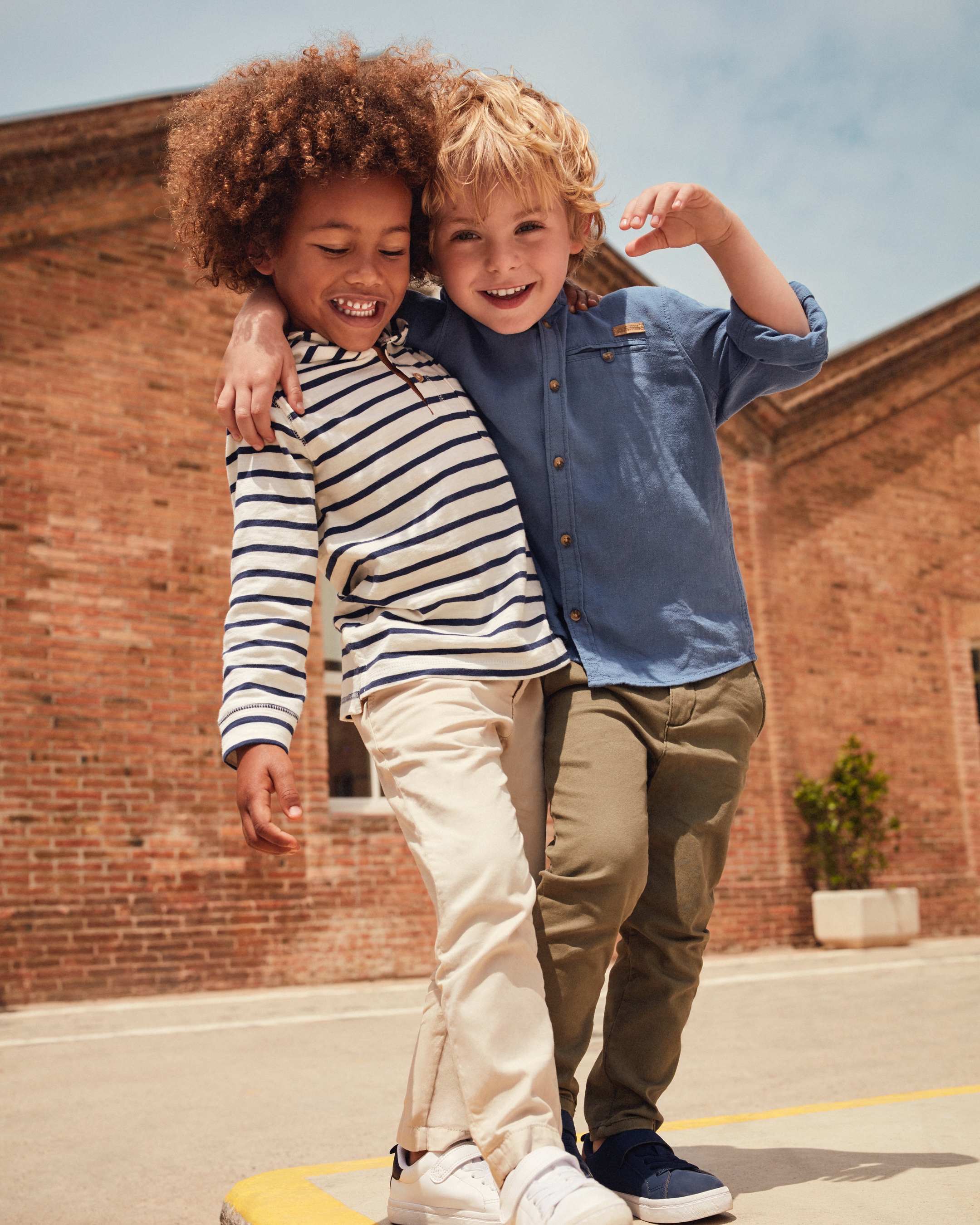 Two boys arms standing, one has his arm over the other boys shoulders, they are smiling. Both are wearing long sleeve shirts and slacks, one wearing white tennis shoes, the other is wearing navy tennis shoes.