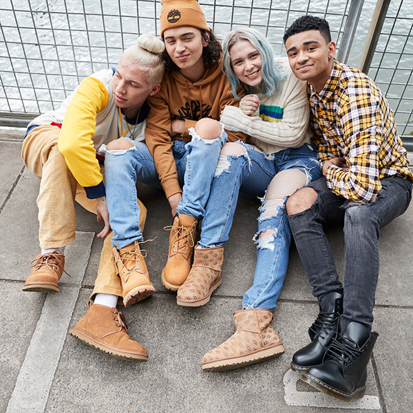 Four teen/young adults sitting in a small group on the ground showing in such a way that you can see their shoes. All wearing a variety of shoes/boots.
