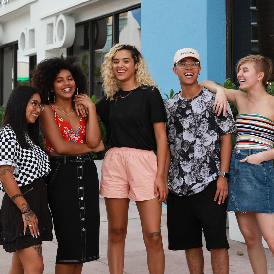 Group of teens/young adults standing together laughing all wearing summer attire, shorts, tank tops, tube tops, skirts.