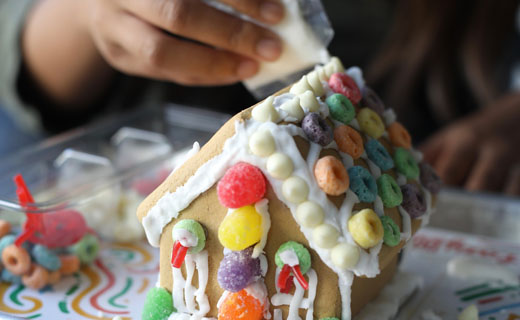 Partially decorated gingerbread house shown with gumdrops and multi-colored fruit cereal being decorated with icing.