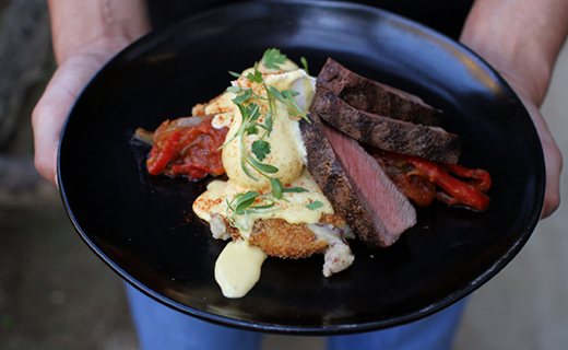 Hands holding a plate with sliced steak, sauteed peppers, poached egg, sauce, and microgreens