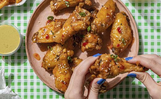 Hands holding chicken wing along with a plate full of chicken wings