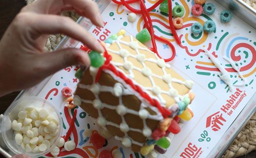 Someone is placing a green gumdrop on a gingerbread house with other candies for decorating shown sitting on a tray with a Habitat for Humanity paper beneath it.