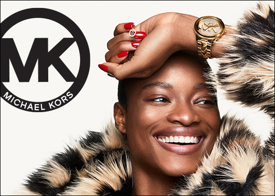 Michael Kors logo. Image: Woman wearing fur coat, she is smiling, her hand is resting on her head showing her red fingernails, gold nail charm and her gold Michael Kors logo watch.