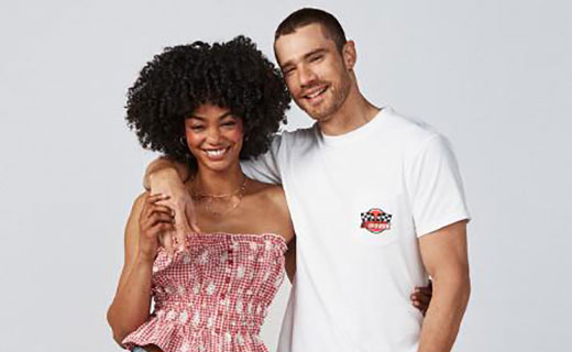 A smiling couple standing close together against a plain background. The woman is wearing a red patterned strapless top and denim shorts, and the man is wearing a white t-shirt and dark shorts with his arm around her shoulders.