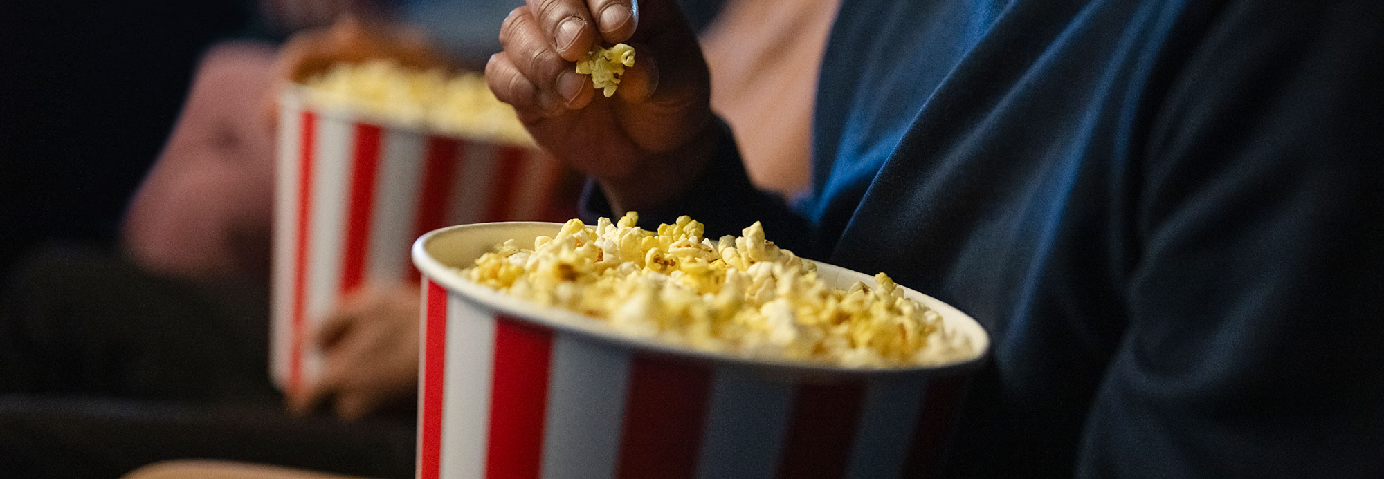 A close-up of a person eating popcorn from a red-and-white striped bucket in a movie theater, with another person holding a similar bucket in the background.