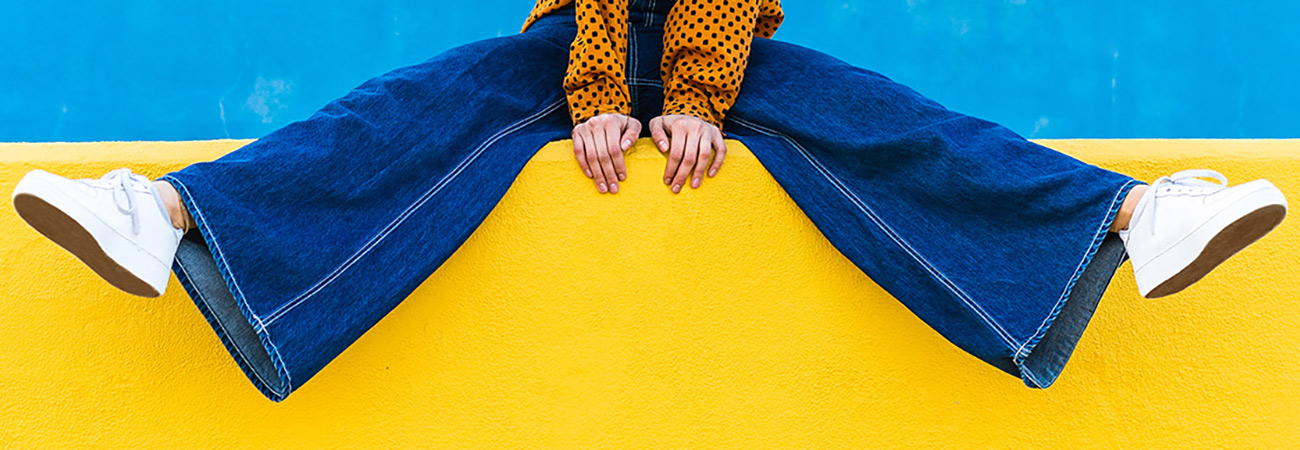 Woman sitting on a yellow wall wearing denim bell-bottom pants, white sneakers and an orange patterned blouse.
