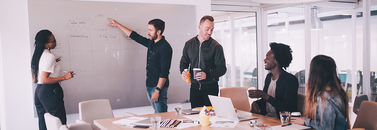 Office meeting room with people sitting at a table and looking at two people present on a whiteboard