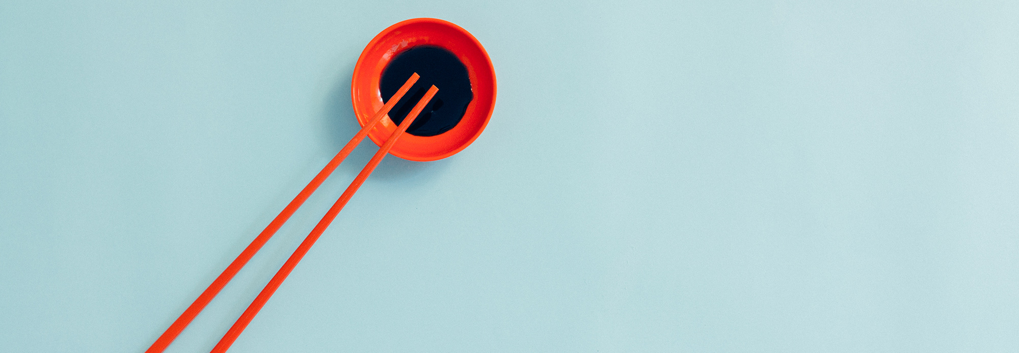 Overhead view of an orange dish of soy sauce and orange chopsticks on a blue background