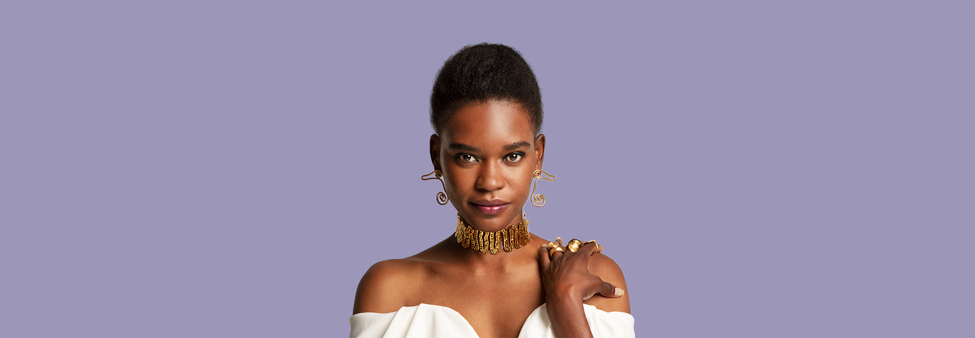 A woman wearing a chunky gold statement choker necklace, statement gold earrings, and a strapless white top.