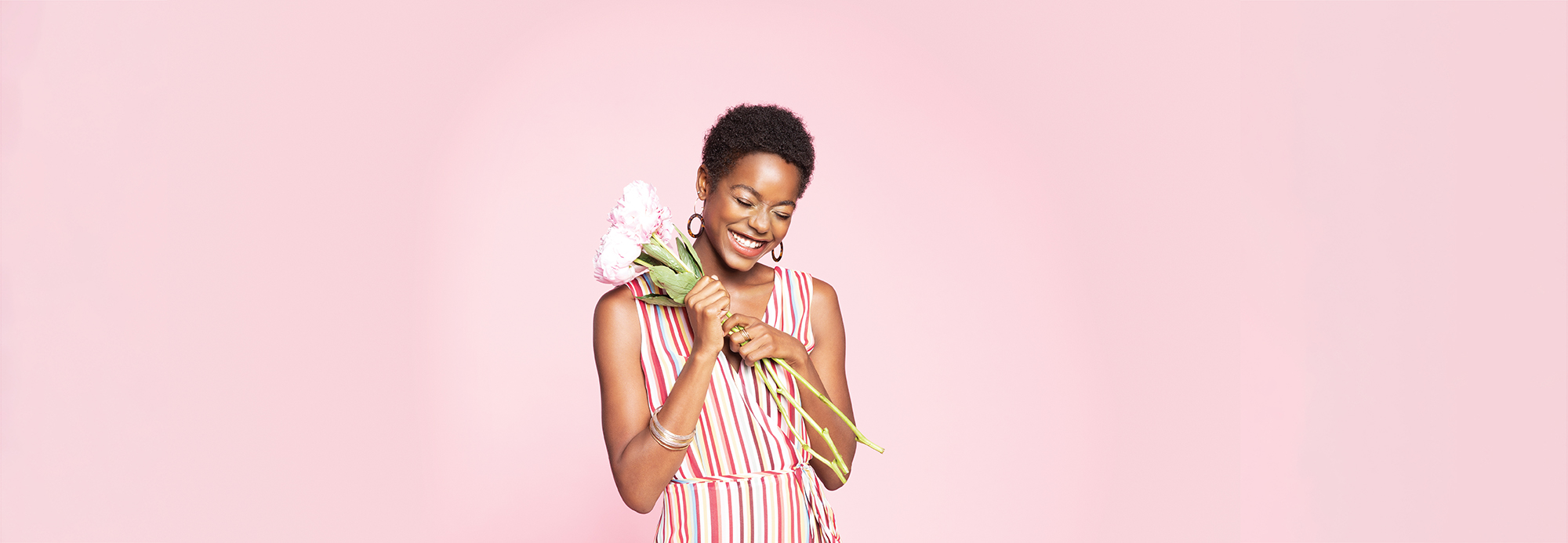 A smiling young woman holding a bouquet and wearing a striped sundress standing in front of a pink background