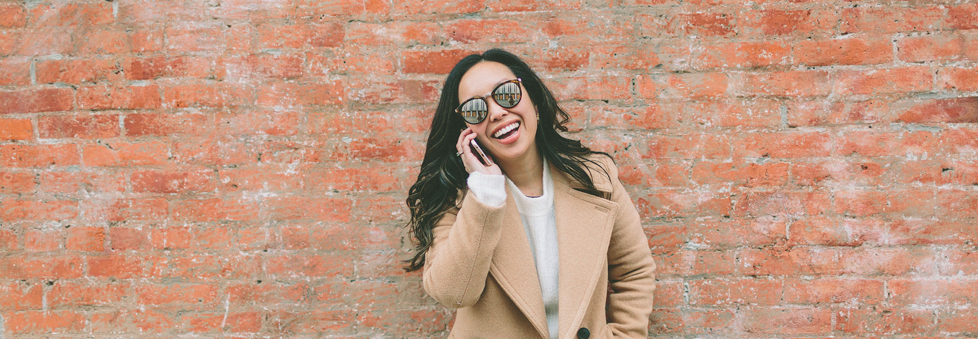 A woman wearing a tan winter coat talking on her cell phone while standing in front of a brick wall