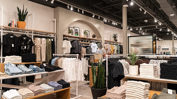 Interior of a modern clothing store featuring neatly folded apparel, hanging racks with neutral-toned clothing, plants throughout the space, bright lighting, and organized retail displays.