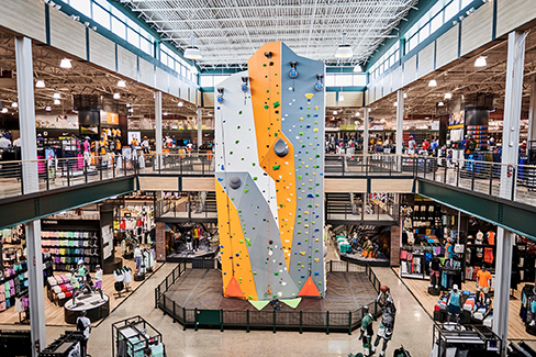 Interior of a large sporting goods store featuring a tall indoor rock climbing wall in the center, multiple retail levels, bright industrial lighting, and shoppers browsing athletic apparel and equipment displays.