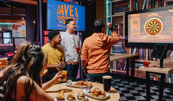 Group of friends playing an interactive dart game inside a Dave & Buster’s entertainment venue, featuring food and drinks on the table, arcade-style lighting, and digital game screens in a lively social setting.