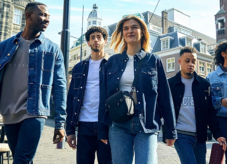 Group of young adults walking outdoors in casual denim and streetwear clothing, smiling and socializing in an urban setting with buildings and sunlight in the background.