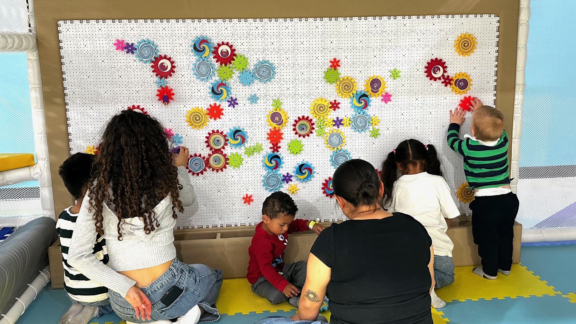 Children and caregivers play together in an indoor family play area, placing colorful gears and spinning shapes on a large interactive wall activity board.