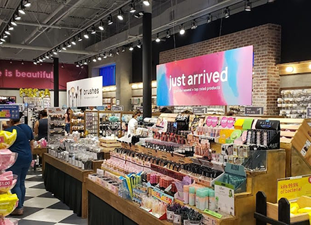 Interior of a beauty and accessories store featuring colorful product displays, makeup and skincare items, bright overhead lighting, wooden shelving, and a large digital sign that reads ‘just arrived.’