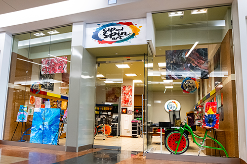 Entrance to Sip & Spin Art studio in a shopping mall, displaying colorful artwork, painting supplies, and a decorative bicycle near the storefront.