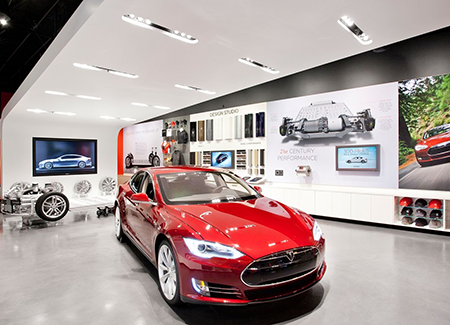 Interior of a Tesla showroom featuring a red Tesla vehicle on display, bright modern lighting, automotive design exhibits on the walls, and interactive displays showcasing electric vehicle technology and performance.