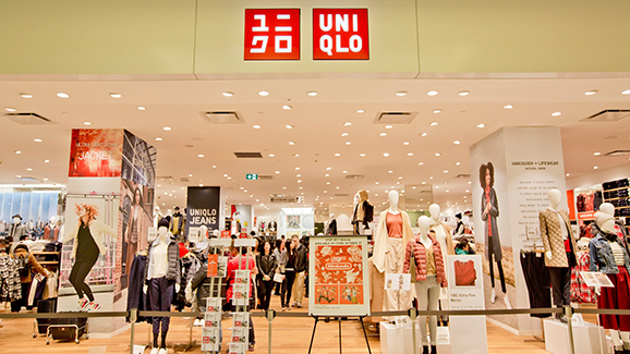 Front entrance of a UNIQLO clothing store featuring bright lighting, mannequins in casual apparel, large red UNIQLO signage, promotional displays, and shoppers browsing modern retail displays inside the store.