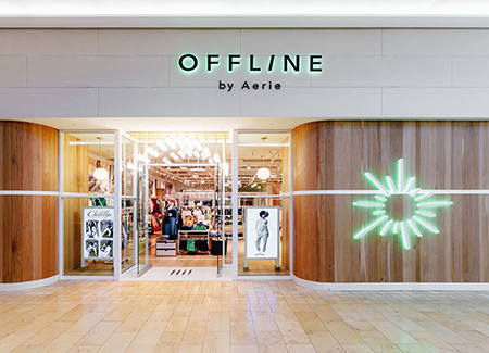 Front entrance of an OFFLINE by Aerie store featuring modern wood-paneled walls, bright interior lighting, activewear displays, large illuminated signage, and a glowing decorative wall light inside a shopping mall.