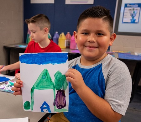 Child holding up a painting he did of a house and tree and clouds with another child in the background painting.