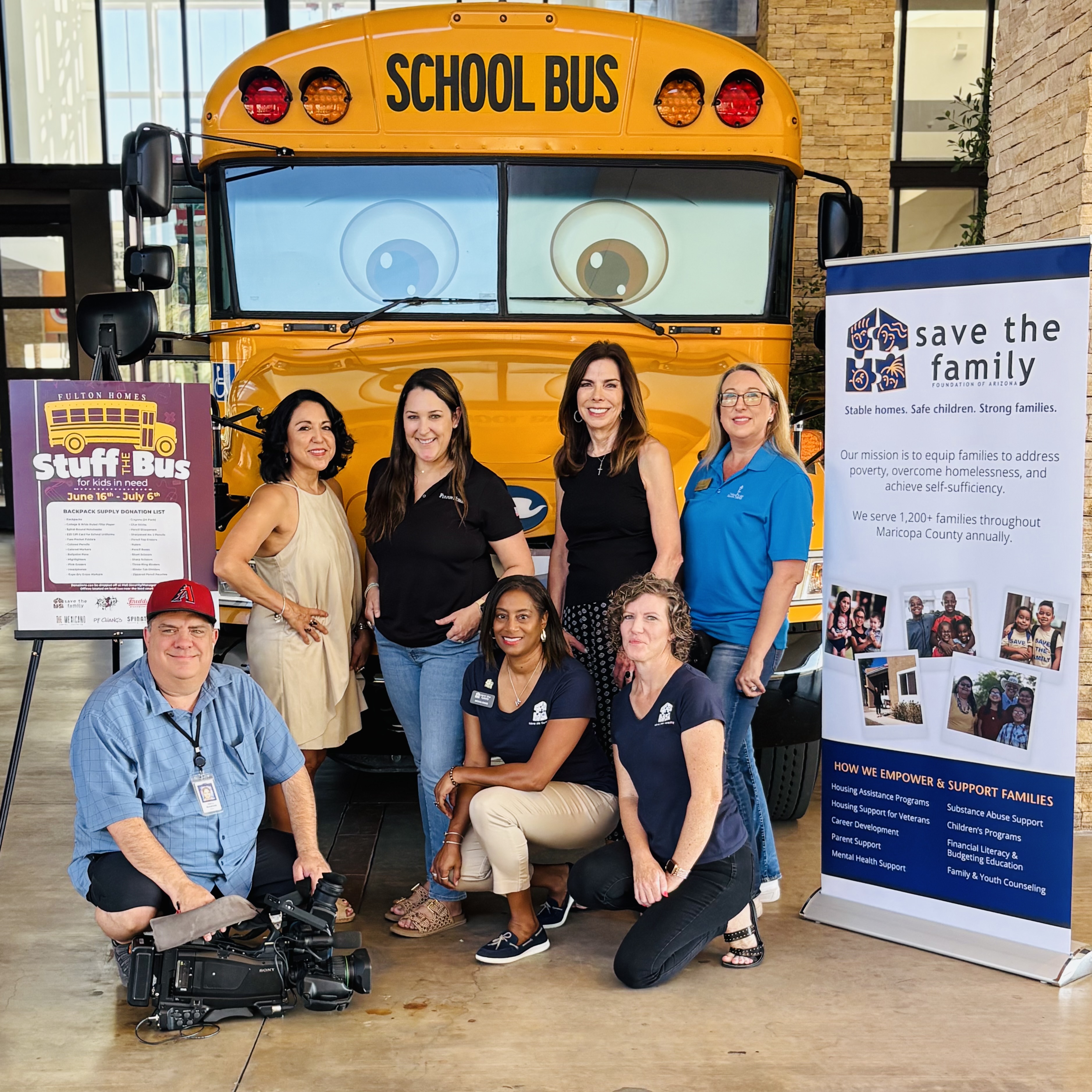 A group of people in front of a school bus that has eyes and a happy face. Standing between two posters, one that says Stuff the Bus, the other says Save the Family.. 