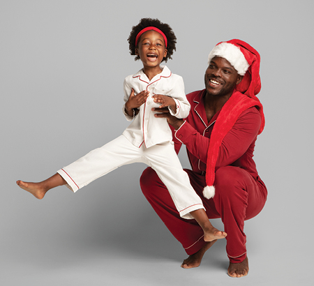 Man dressed in red pajamas wearing a Santa hat holding a little girl in white pajamas