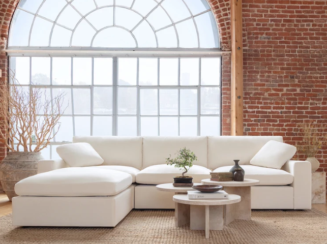 Modern white sectional sofa in a bright loft-style living room with exposed brick walls, a large arched window, round coffee tables, and minimalist decor.