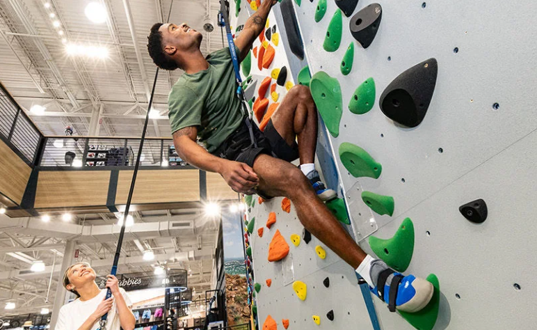 Person wearing a harness climbs an indoor rock wall with colorful handholds while another person below manages the safety rope in a brightly lit recreation facility.