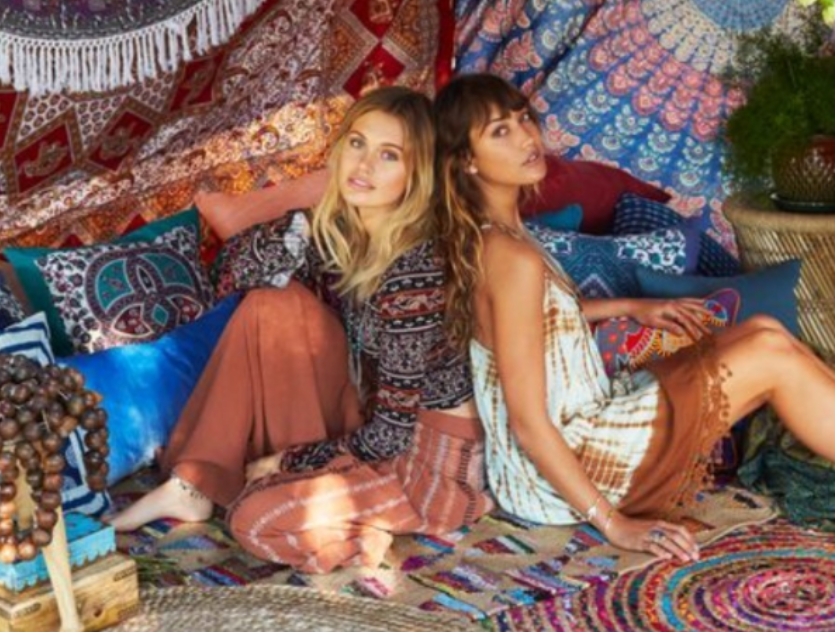 Two people sitting back-to-back in a colorful, bohemian-style tent decorated with patterned rugs, pillows, and tapestries.